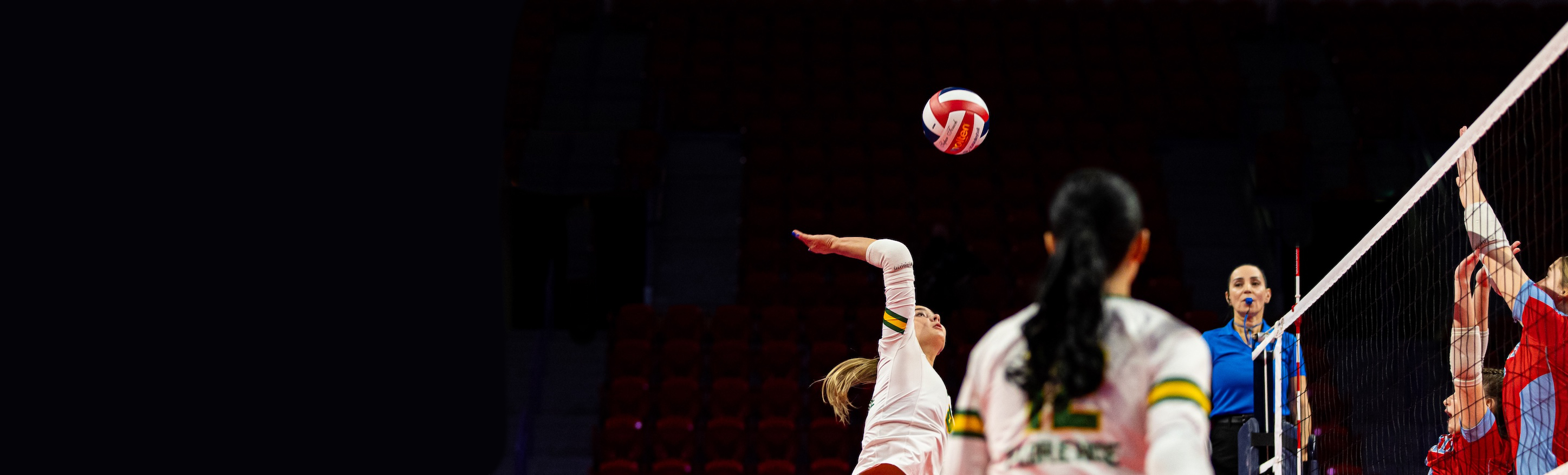 A volleyball player in a white uniform jumps to attack the ball near the net during an indoor match, while two opposing blockers reach up to defend. Teammates watch the play unfold as the ball travels above the court under arena lighting.