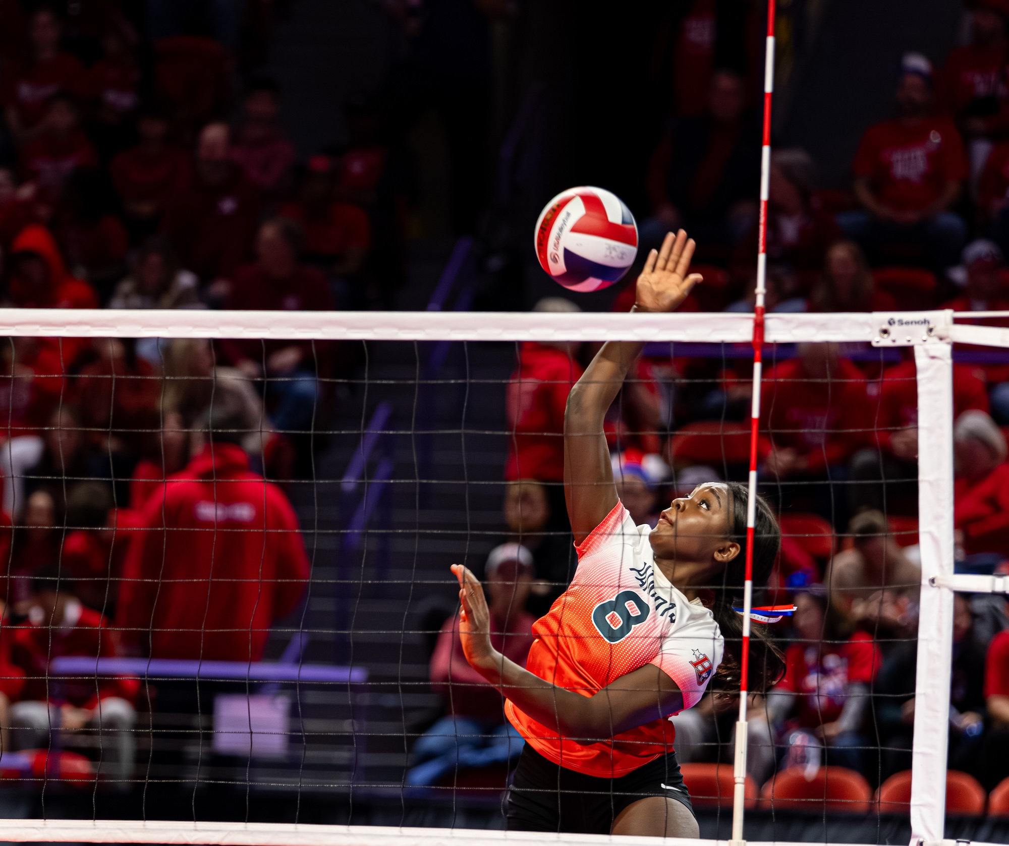 A volleyball player in an orange and white uniform reaches above the net to tip the ball during an indoor match. With eyes locked on the ball and one hand extended, the player redirects the attack as spectators watch from the stands behind the court.