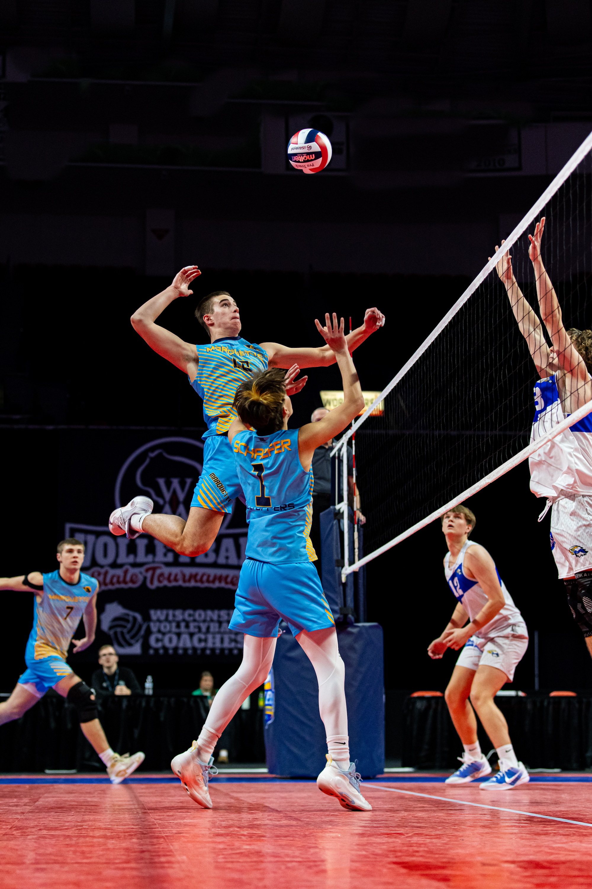 High school boys competing in an indoor volleyball match during a state tournament, with a player jumping to spike the ball near the net as teammates and defenders prepare for the play.