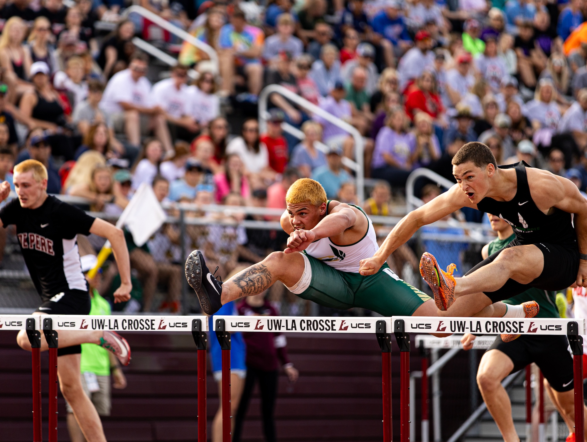 Athletes leap over hurdles during a crowded track-and-field race, with two runners captured midair in an intense side-by-side sprint.