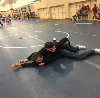 Two people demonstrate a wrestling hold on a mat, with one person controlling the other’s upper body from a side position.