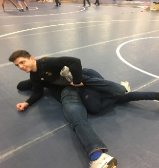 Two people lie on a wrestling mat during a grappling drill, with one person positioned partly across the other’s torso.