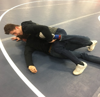 Two people practice a wrestling hold on a mat, with one person lying on their back while the other maintains control from the side.