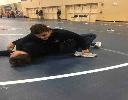 Two people pause during a wrestling practice drill, with one person leaning over the other on the mat.