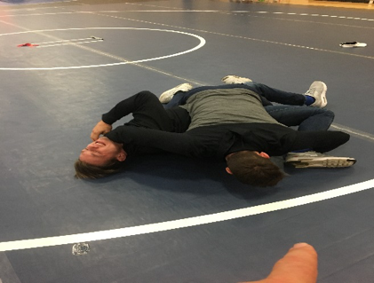 Two people practice a wrestling control position, with one person lying face down while the other maintains pressure from the side.