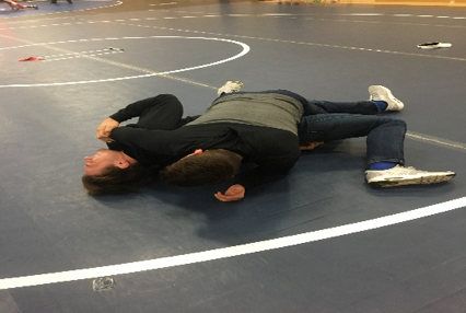 Two people lie on a wrestling mat during a grappling drill, with one person positioned partly across the other’s torso.