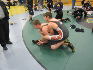 Two wrestlers practice a mat drill, with one wrestler positioned behind the other and controlling their leg.