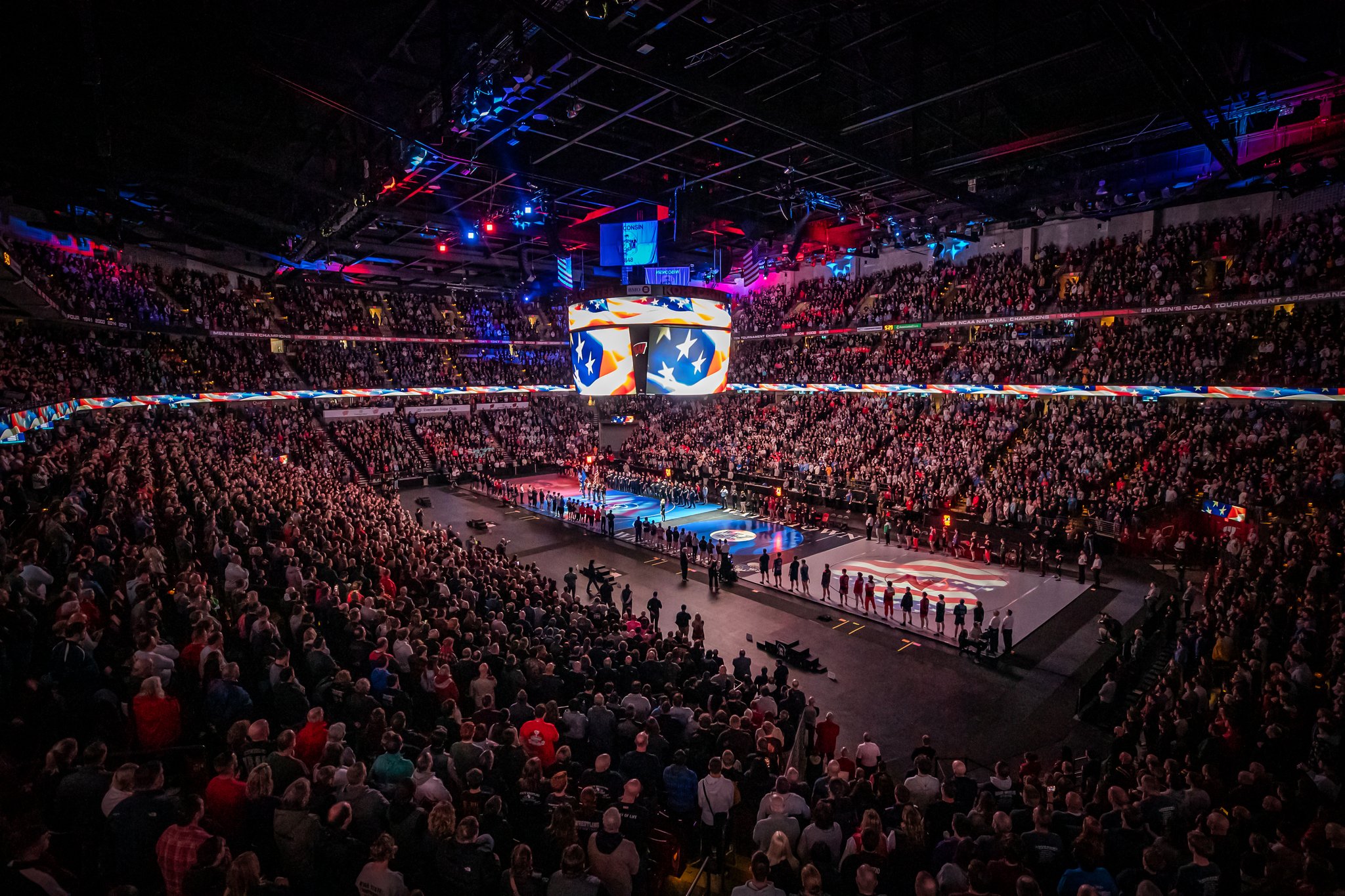 Wide view of a packed arena during a wrestling championship event, with athletes lined up around the mats under colorful lights and large video displays.