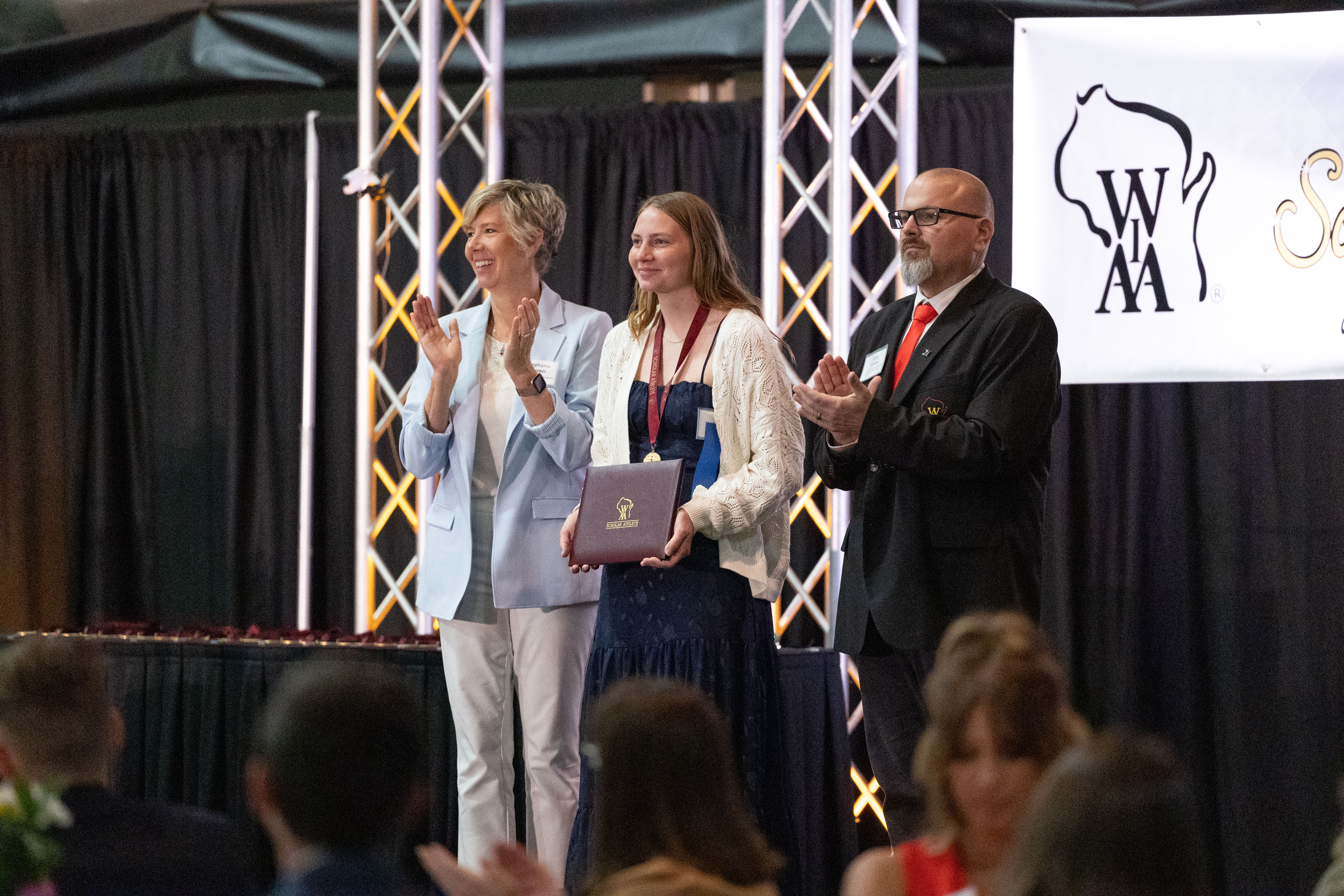 Student wearing a medal holds an award plaque on stage while two adults applaud during a WIAA ceremony.