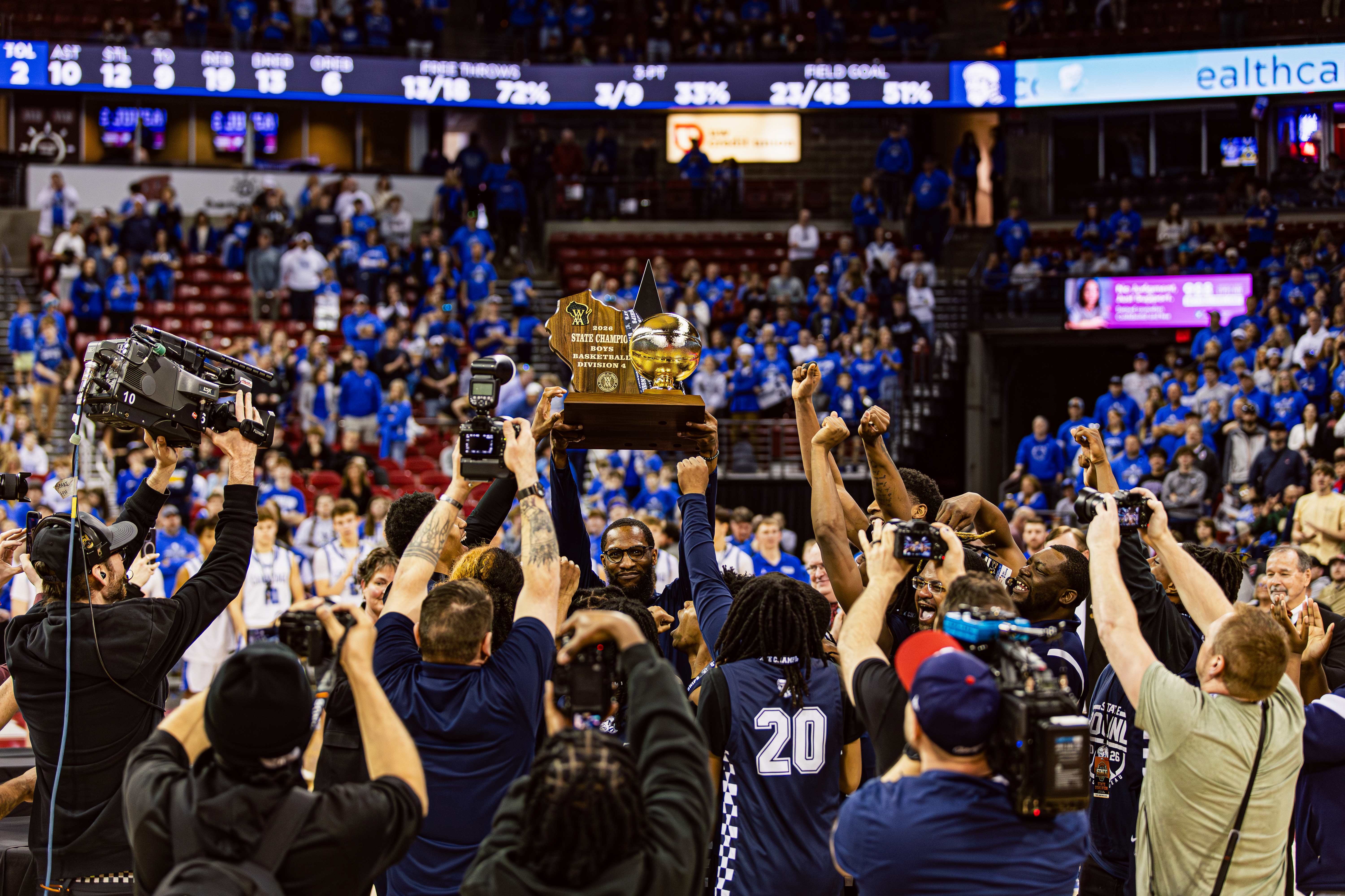 Basketball team celebrates by lifting a 2026 State Champion Boys Basketball Division 4 trophy in a crowded arena as cameras surround them.