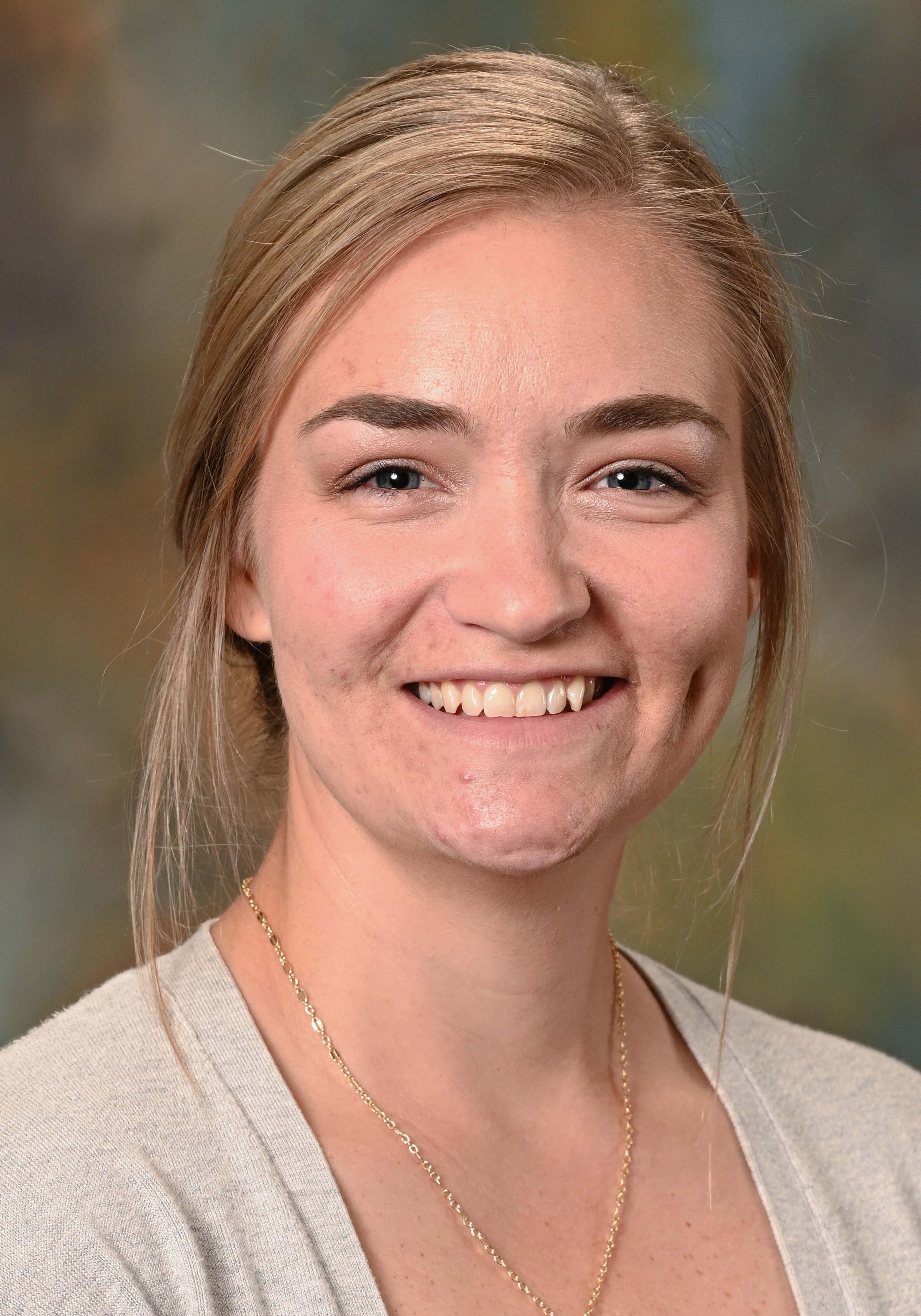 Headshot of a smiling woman with blonde hair pulled back, wearing a light gray cardigan and gold necklace.