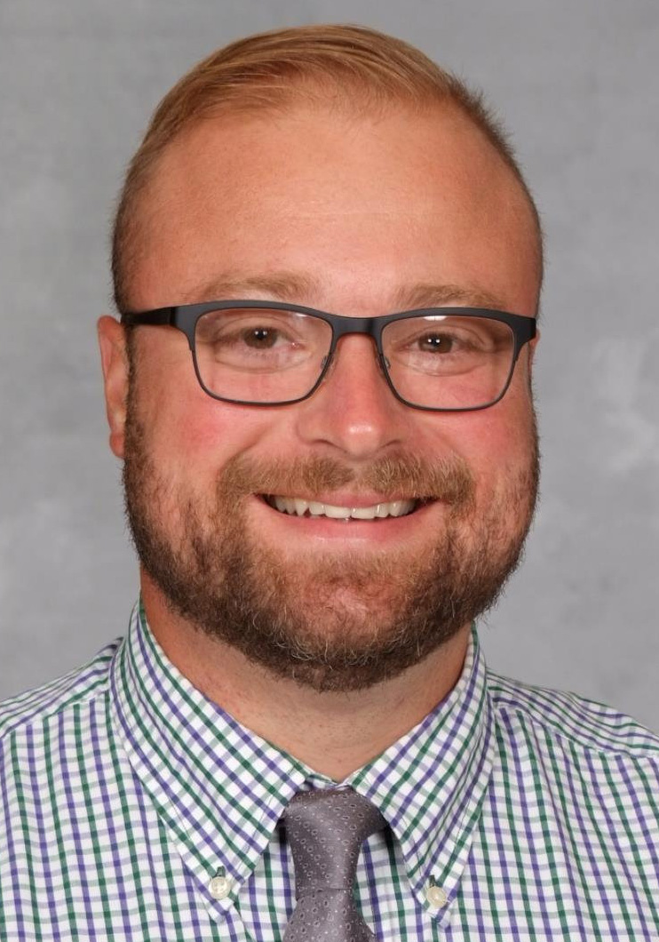 Headshot of a smiling man wearing glasses, a green and purple checkered shirt, and a gray tie against a gray background.
