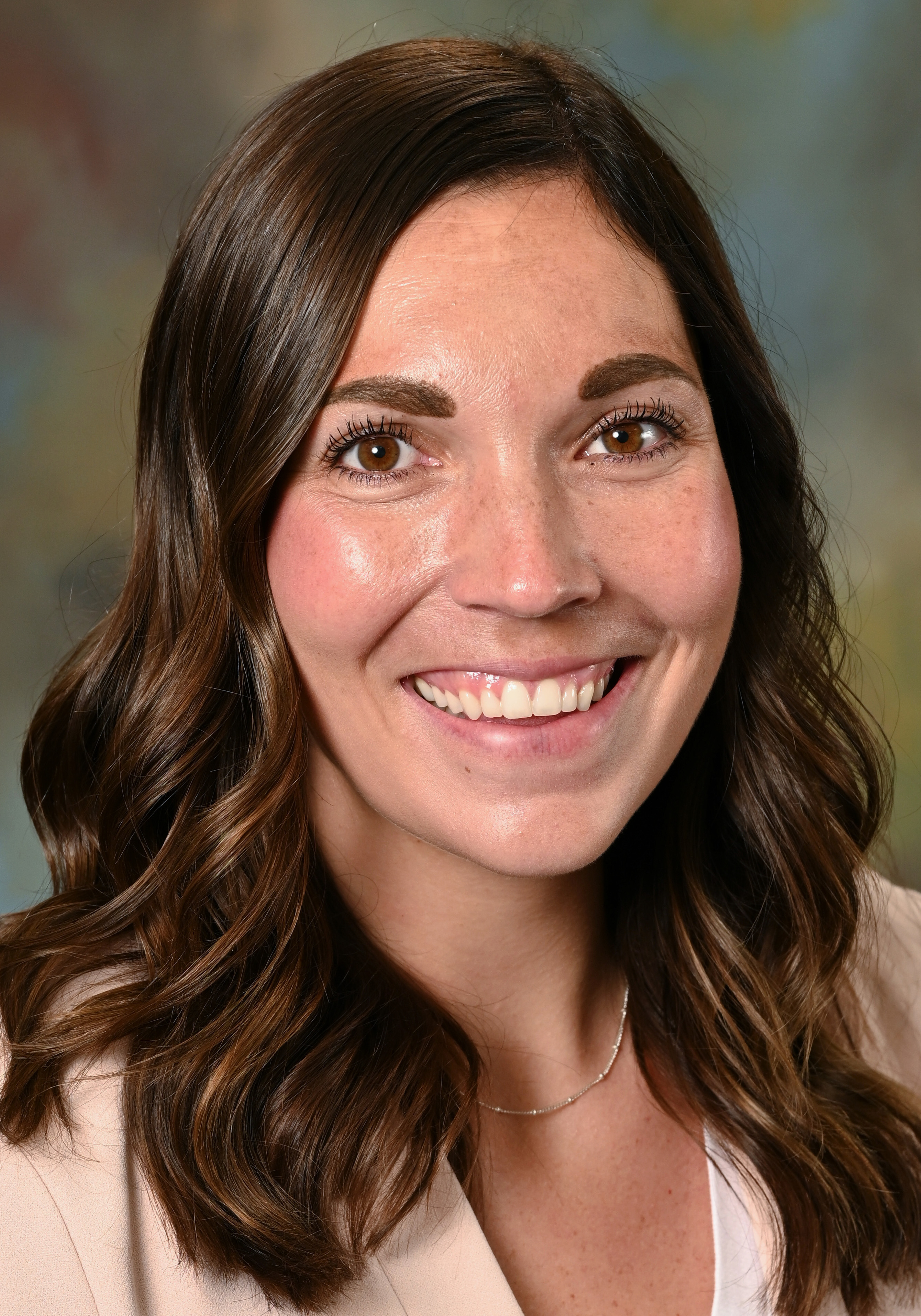 Headshot of a smiling woman with long wavy brown hair wearing a light blazer and white top.