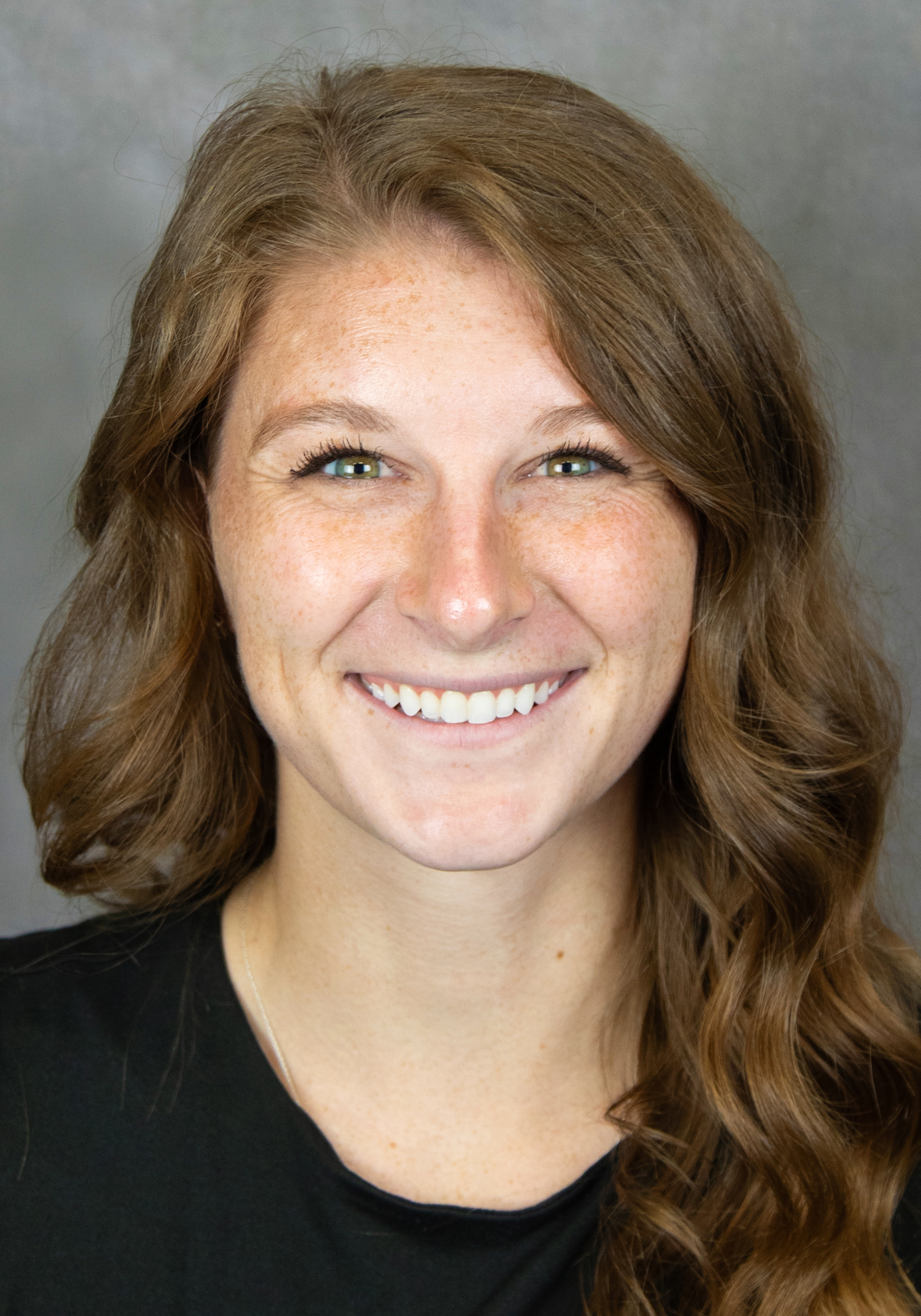 Headshot of a smiling woman with long wavy brown hair wearing a black top against a gray background.