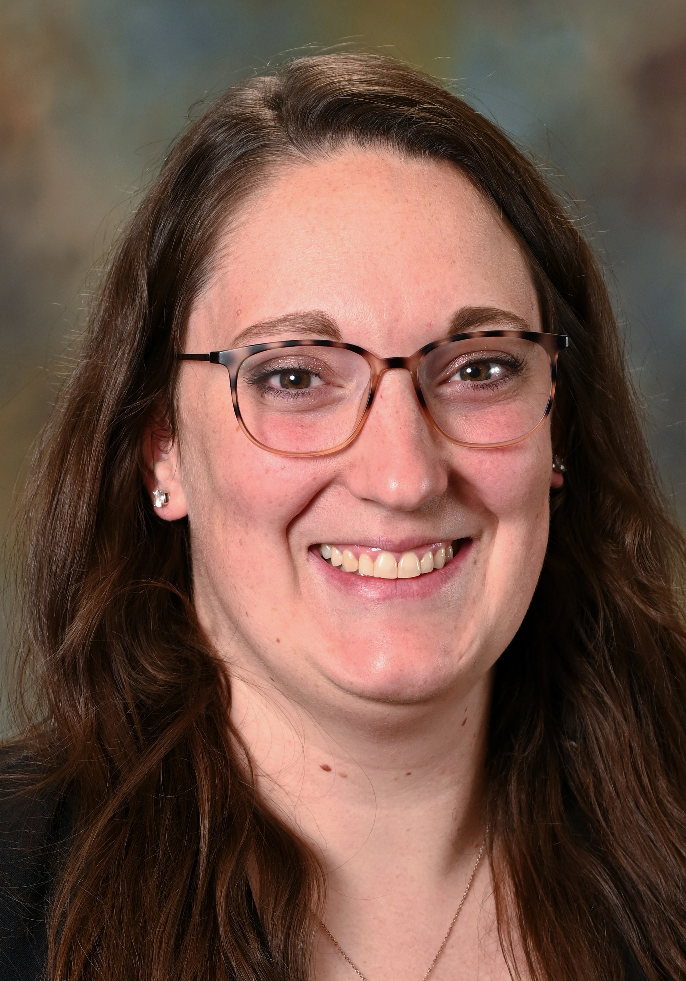 Headshot of a smiling woman with long brown hair and glasses wearing a black blazer.