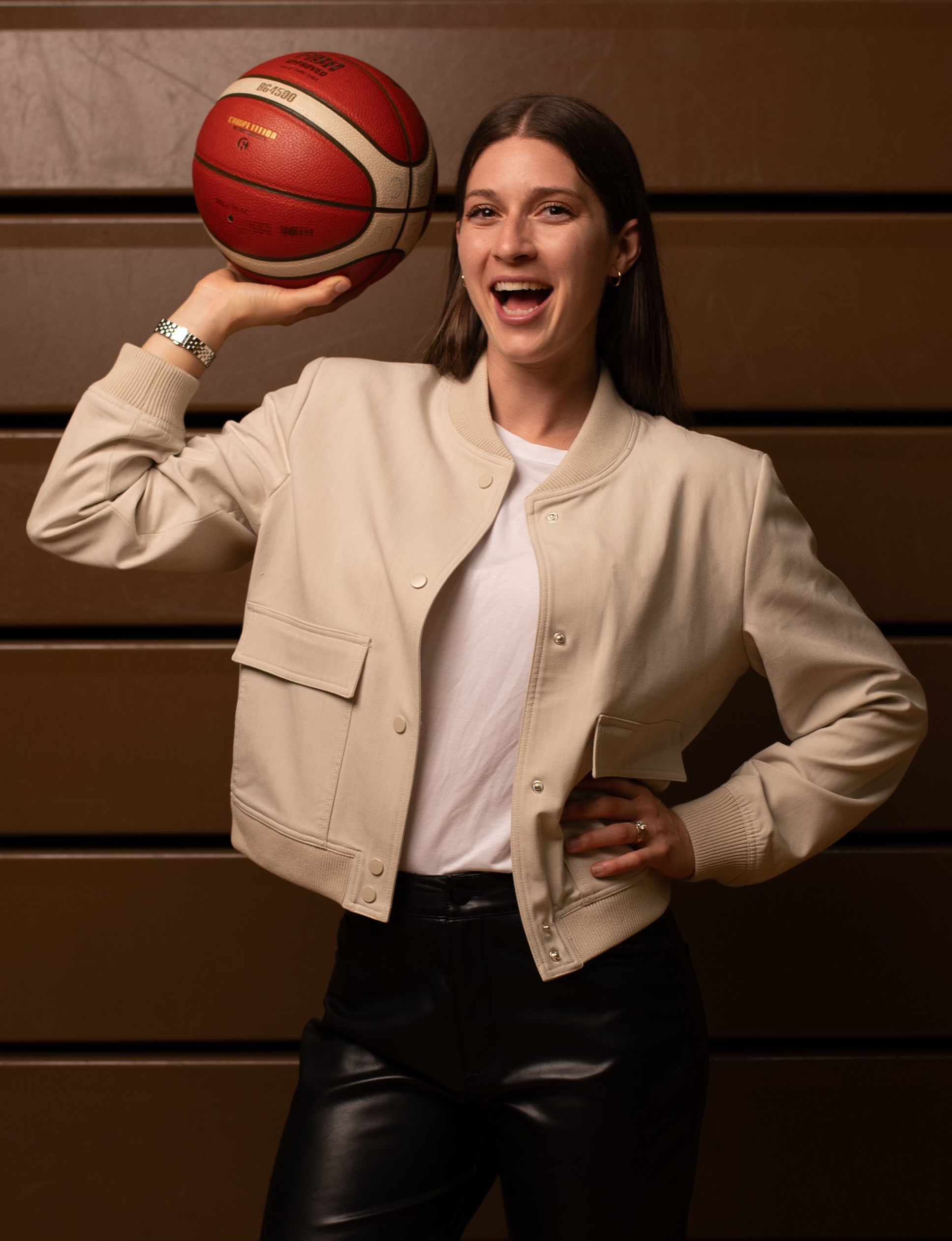 A smiling woman poses with one hand on her hip while holding a basketball near her shoulder.