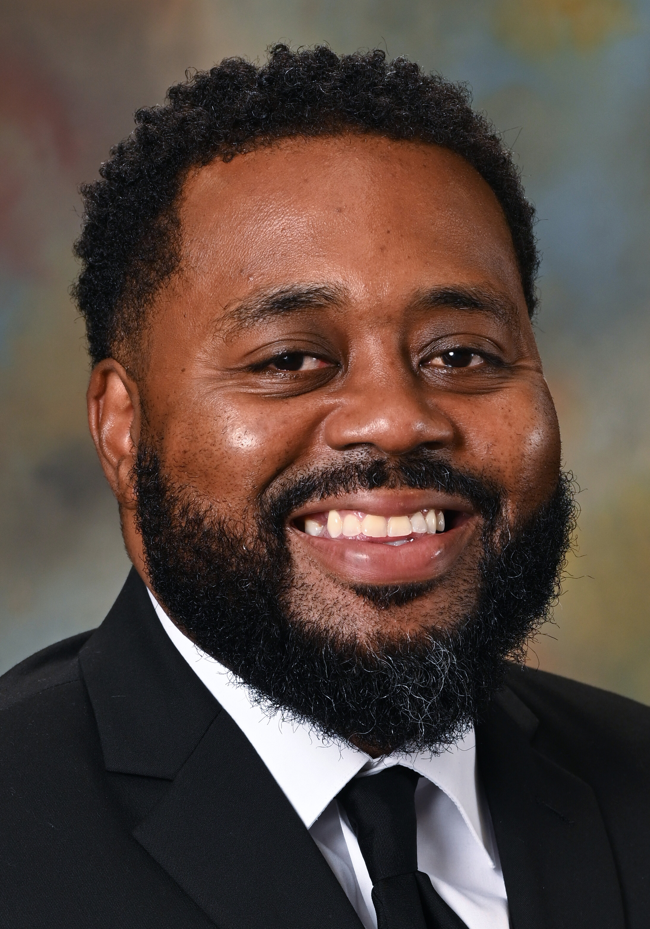 Headshot of a smiling man with short curly hair and a beard wearing a black suit, white shirt, and black tie.