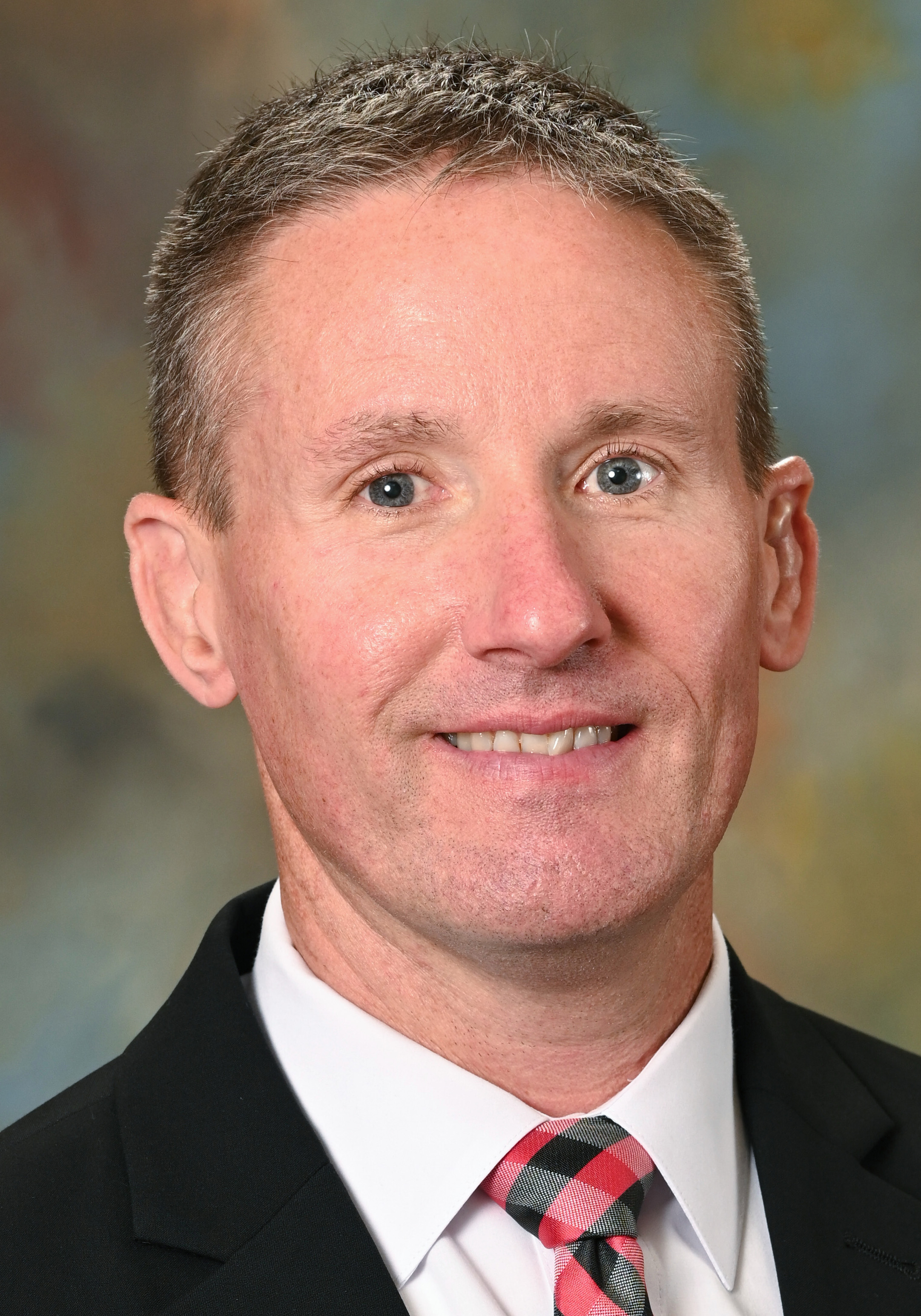 Man in a suit and red plaid tie posing for a professional headshot.