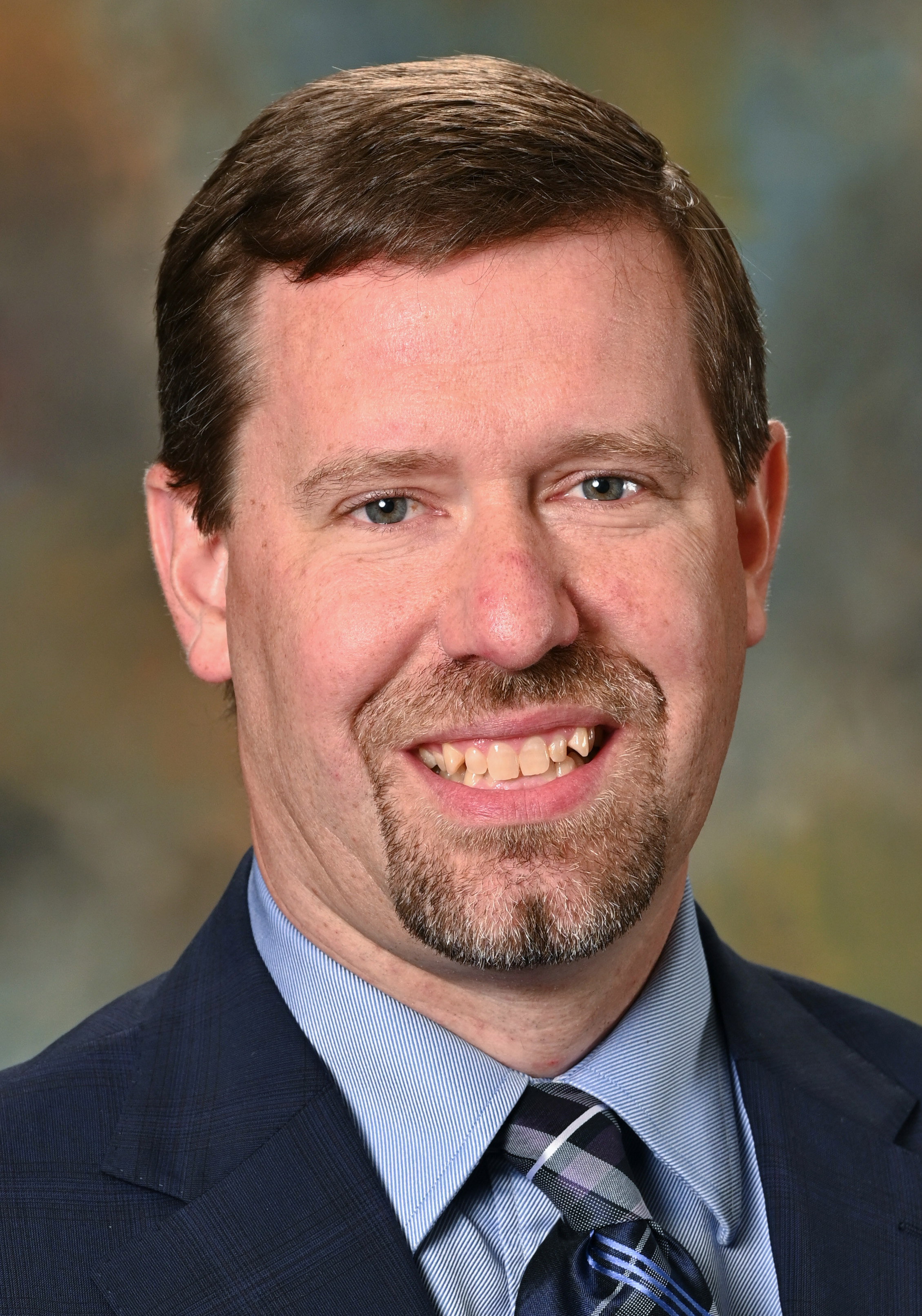 Headshot of a smiling man with short brown hair and a goatee wearing a navy suit, blue shirt, and patterned tie.