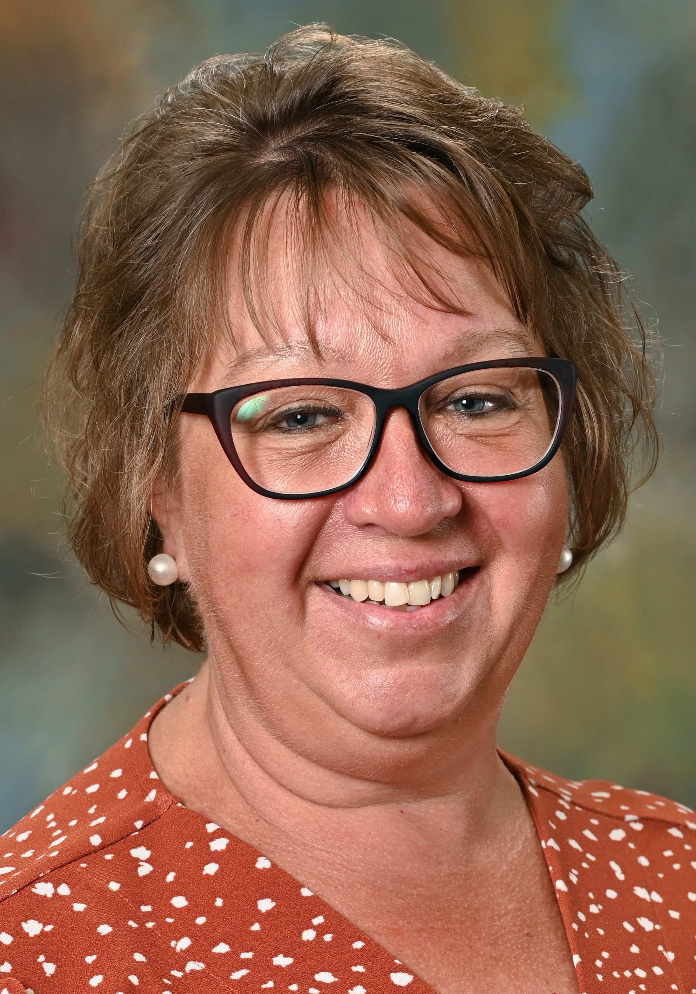 Headshot of a smiling woman with glasses wearing pearl earrings and an orange patterned blouse.