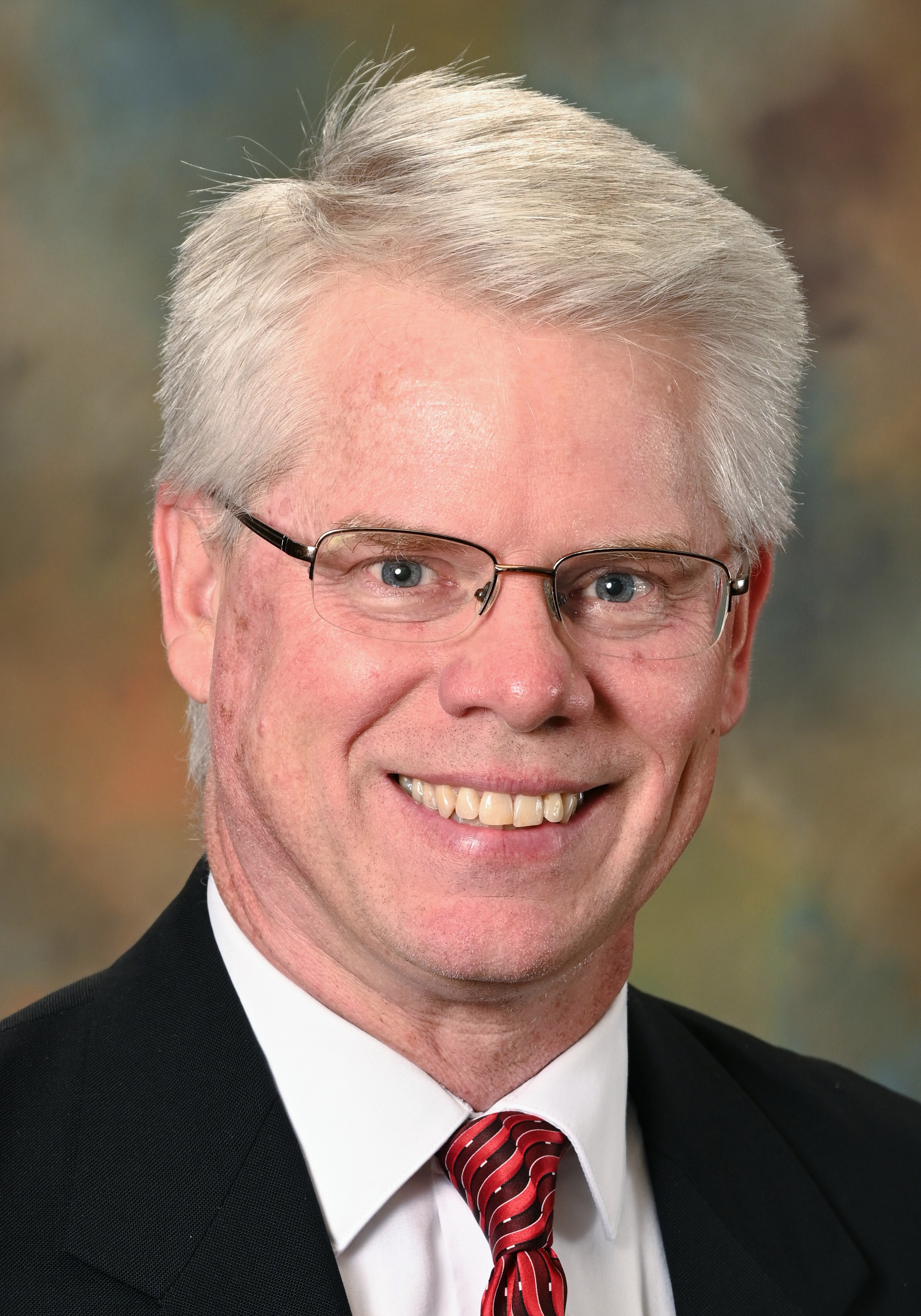 Headshot of a smiling man with gray hair and glasses wearing a black suit, white shirt, and red striped tie.