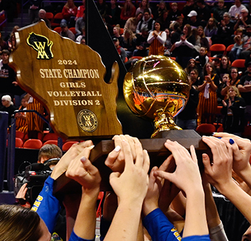 Players hold up a 2024 State Champion Girls Volleyball Division 2 trophy and gold volleyball award in front of a cheering crowd.