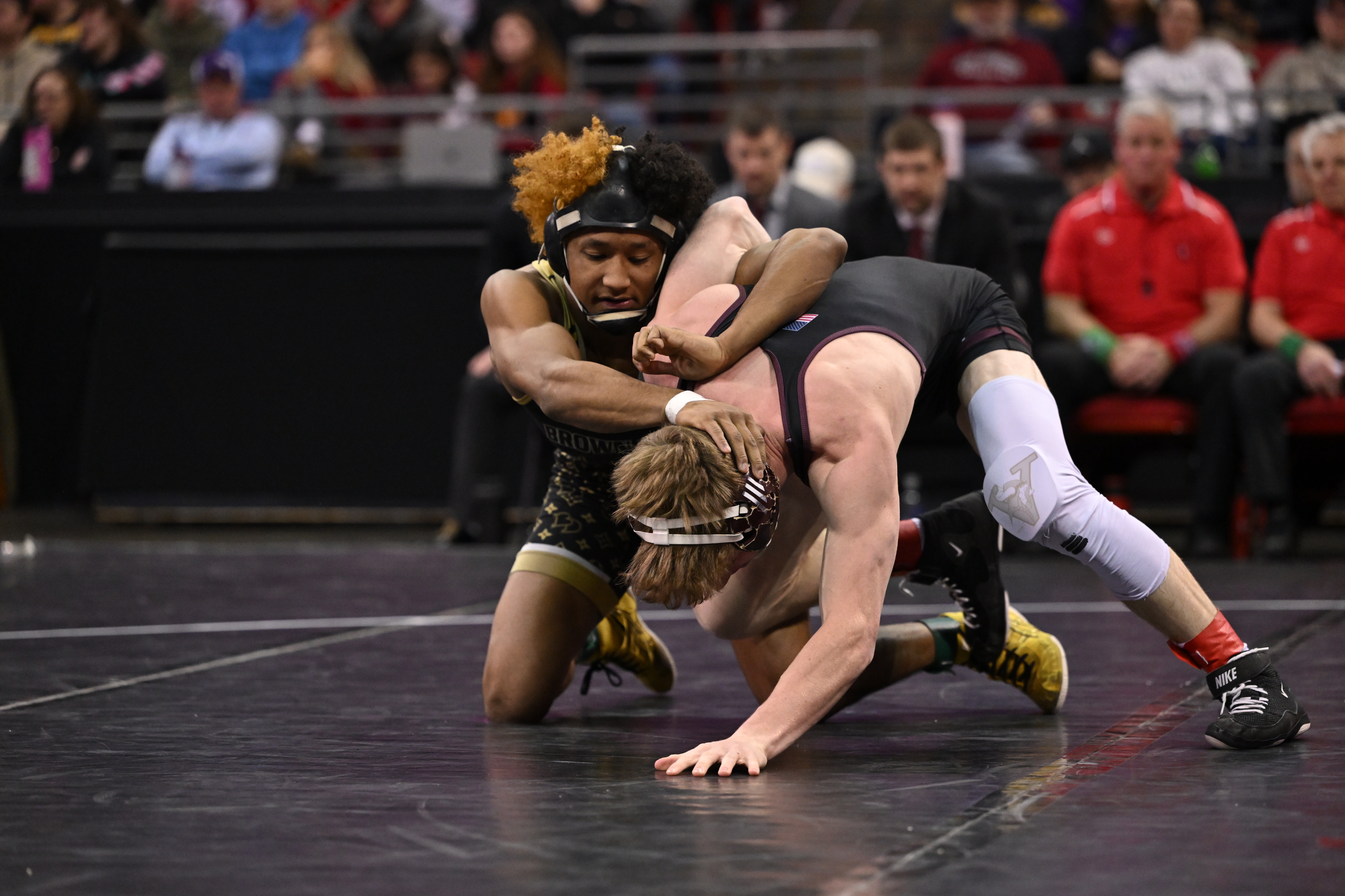 Two wrestlers compete on the mat during a match, with one athlete controlling the other in a front headlock position as spectators and officials watch in the background.