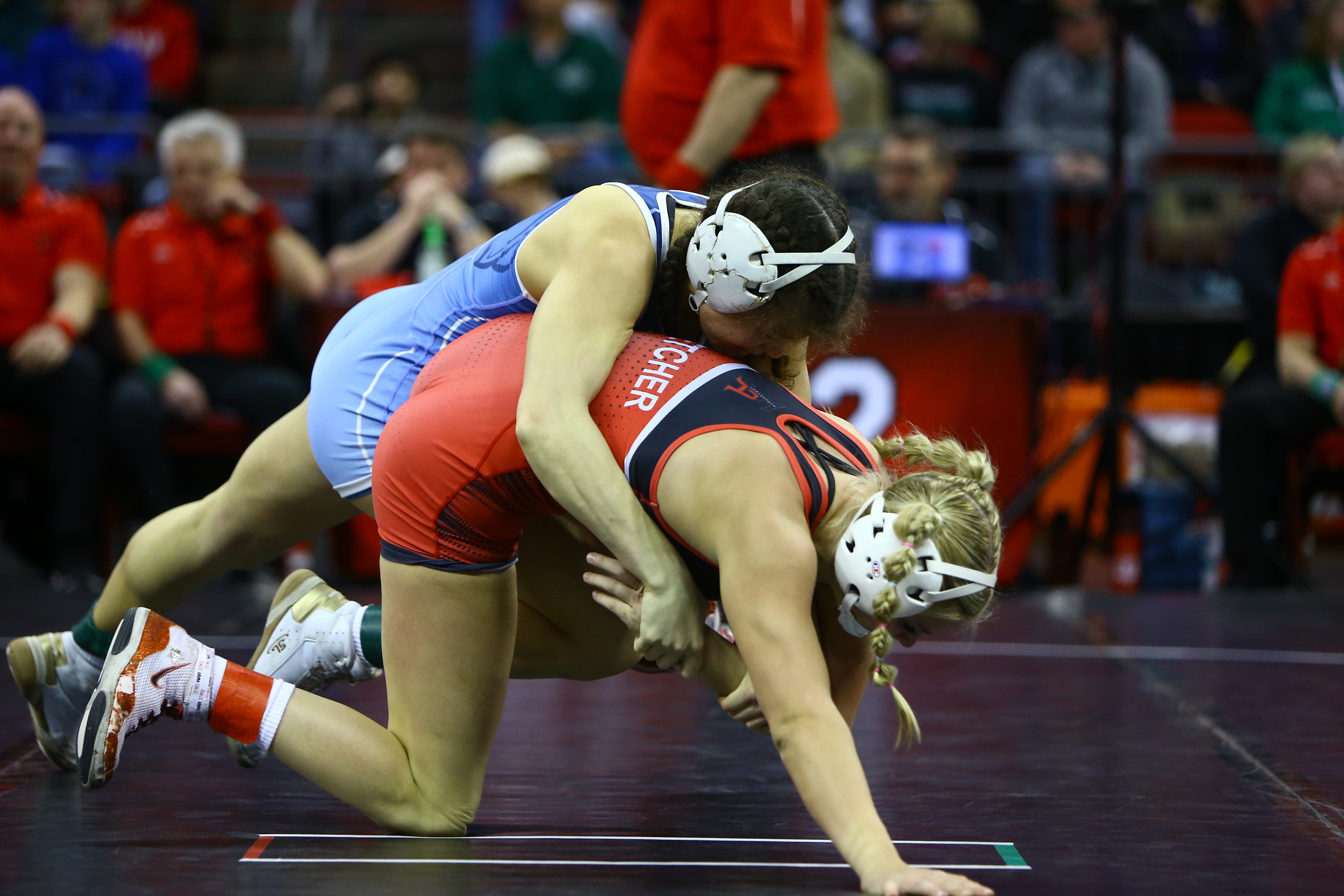 Two wrestlers compete on the mat, with one athlete in blue controlling an opponent in red from behind during a match.
