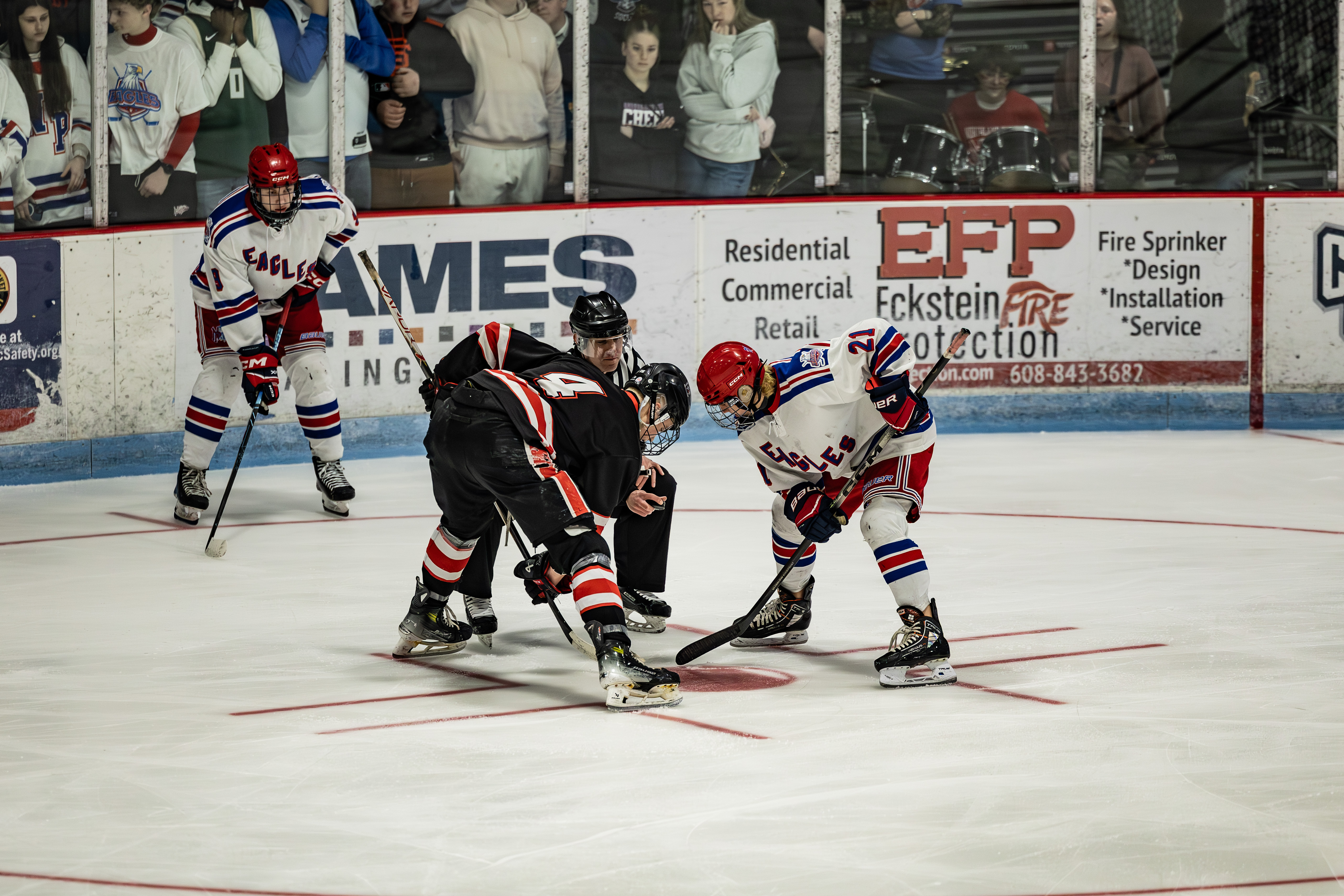 Two ice hockey players face off at center ice during a game, with teammates and spectators watching from behind the boards.
