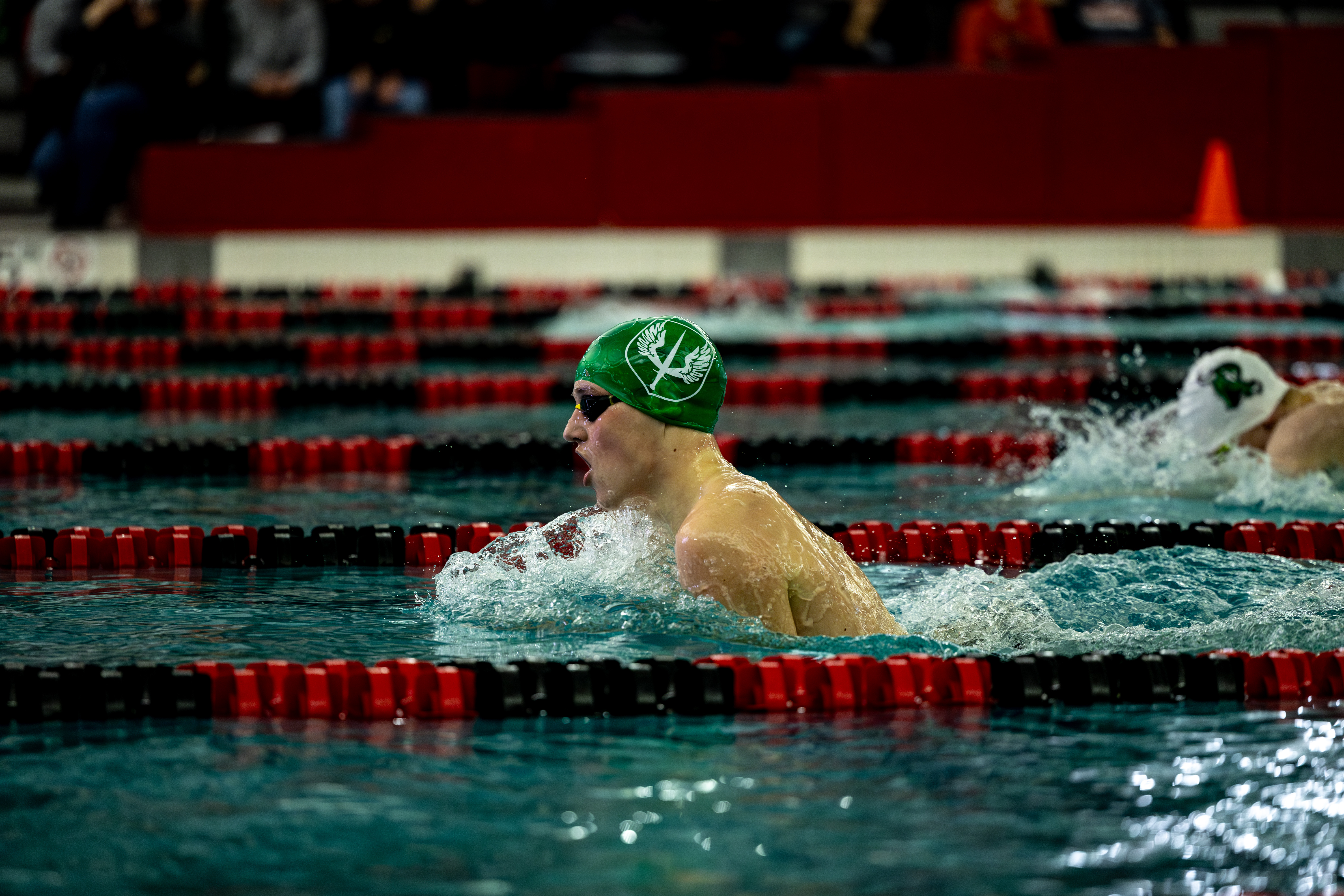 A swimmer in a green cap competes in an indoor breaststroke race, moving through a lane with water splashing around them.