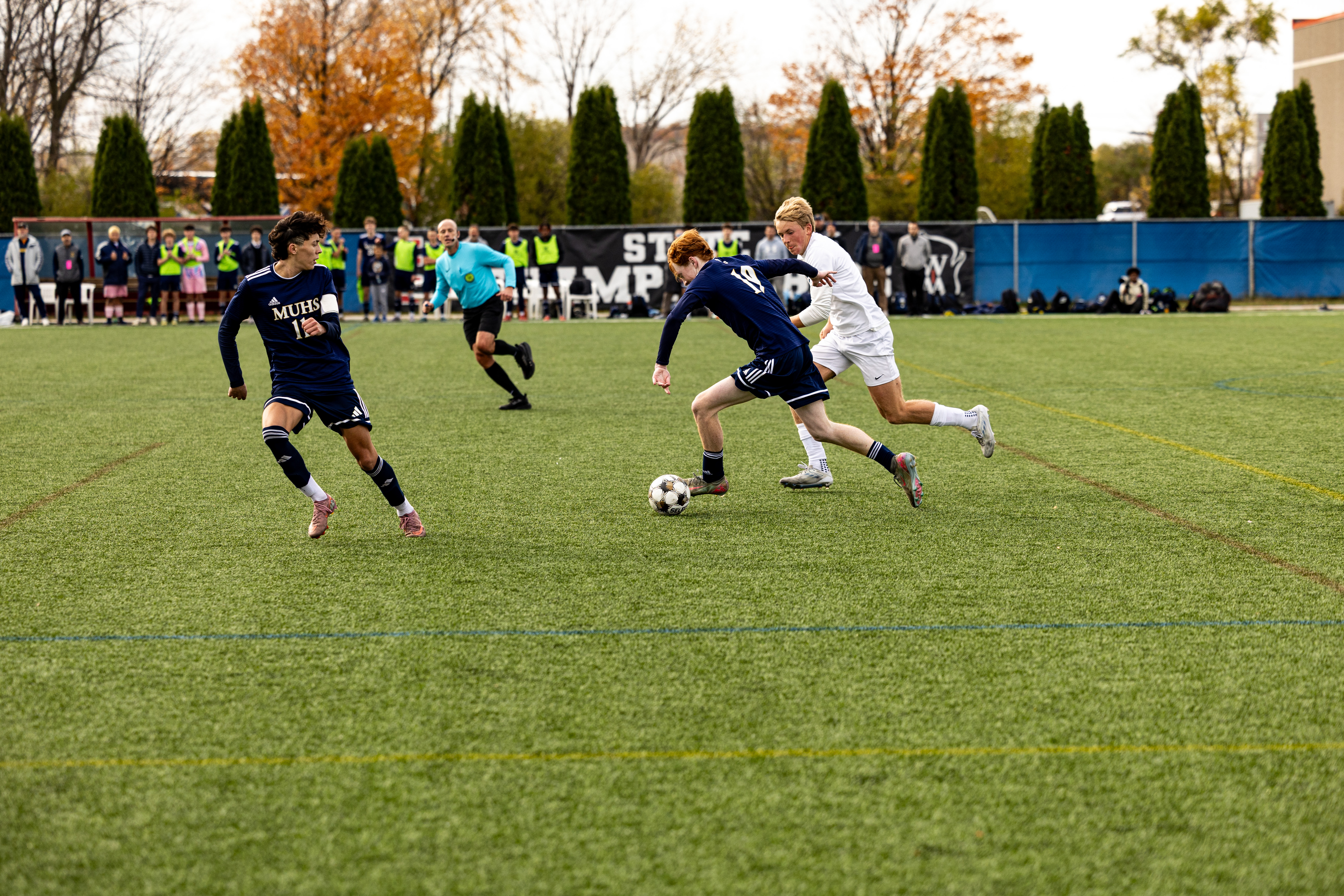 Soccer players compete for possession of the ball during an outdoor game, with a referee and spectators in the background.
