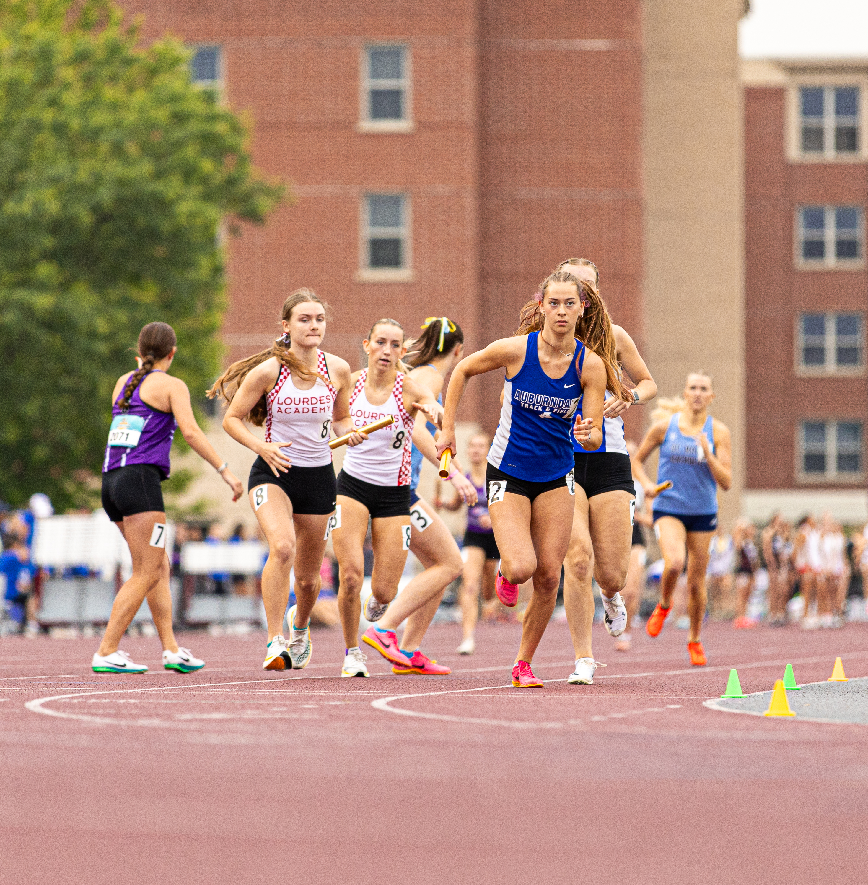 High school girls compete in a track relay race, with runners carrying batons around a curve on an outdoor track.