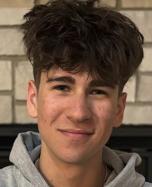 Young man with dark wavy hair smiling slightly while standing in front of a brick wall.