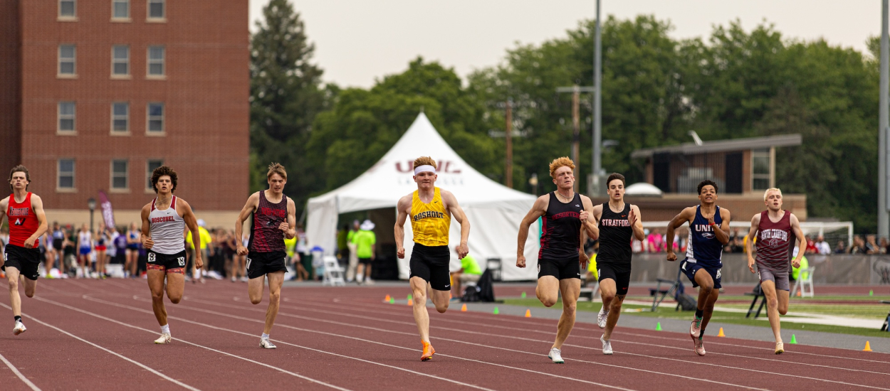 High school boys competing in a sprint race on an outdoor track during a championship meet, with athletes spread across lanes as they race toward the finish line.