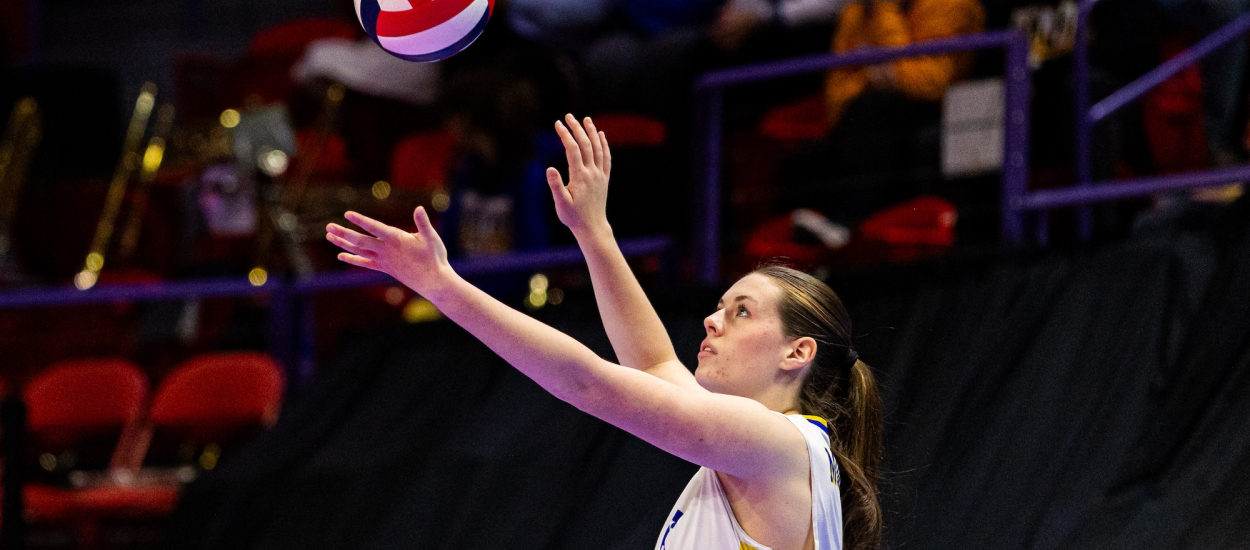 A volleyball player in a white, blue, and yellow uniform prepares to serve the ball during an indoor match. With arms raised and eyes focused upward, the player tracks the ball toss as spectators watch from the stands in the background.