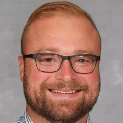 Headshot of a smiling man wearing glasses, a green and purple checkered shirt, and a gray tie against a gray background.