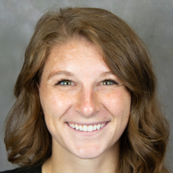 Headshot of a smiling woman with long wavy brown hair wearing a black top against a gray background.