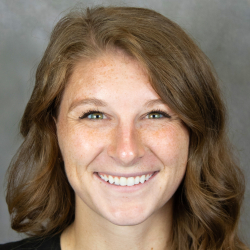 Headshot of a smiling woman with long wavy brown hair wearing a black top against a gray background.