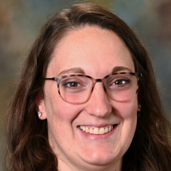 Headshot of a smiling woman with long brown hair and glasses wearing a black blazer.