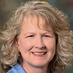 Headshot of a smiling woman with curly blonde hair wearing a light blue collared shirt.