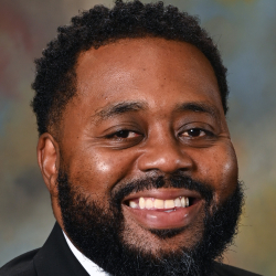 Headshot of a smiling man with short curly hair and a beard wearing a black suit, white shirt, and black tie.