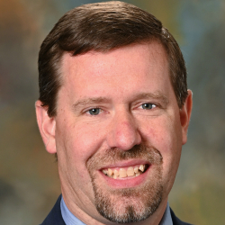 Headshot of a smiling man with short brown hair and a goatee wearing a navy suit, blue shirt, and patterned tie.