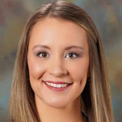 Headshot of a smiling woman with long highlighted hair wearing a black blazer, white top, and necklace.