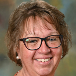 Headshot of a smiling woman with glasses wearing pearl earrings and an orange patterned blouse.