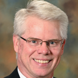 Headshot of a smiling man with gray hair and glasses wearing a black suit, white shirt, and red striped tie.